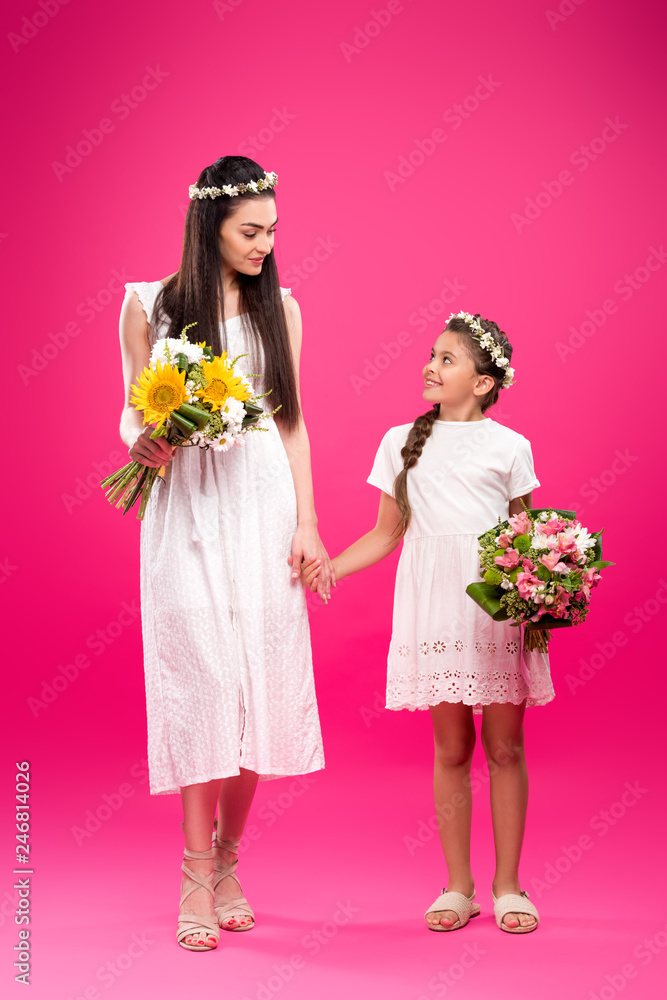 full length view of beautiful mother and daughter in white dresses holding bouquets and smiling each other on pink full length view of beautiful mother and daughter in white dresses holding bouquets and smiling each other on pink