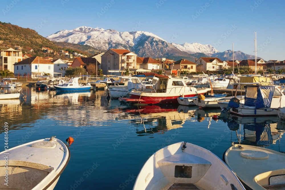 Sunny winter day. Fishing boats in harbor in  Mediterranean town at foot of snowy mountains. Montenegro, Bay of Kotor, Marina Kalimanj in Tivat city and snow-capped of Lovcen mountains