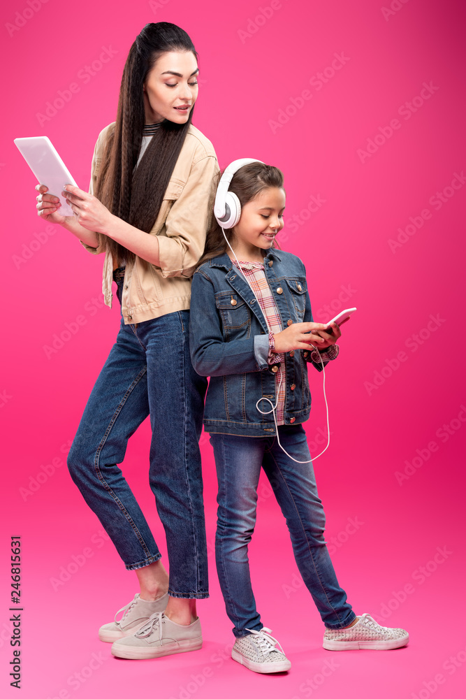 Fototapeta premium full length view of smiling mother holding digital tablet and looking at daughter in headphones using smartphone on pink