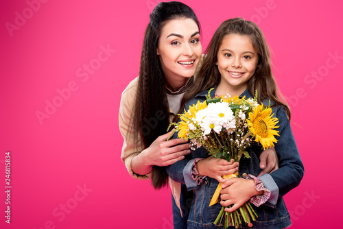 happy mother and daughter holding beautiful bouquet of flowers and smiling at camera isolated on pink