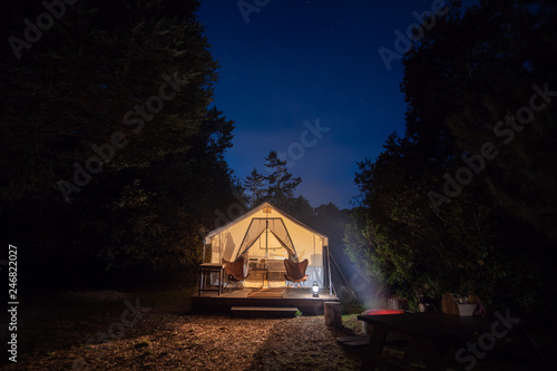 Camping tent site with a camp fire under the blue night sky with an illuminated tent and trees in the background.
