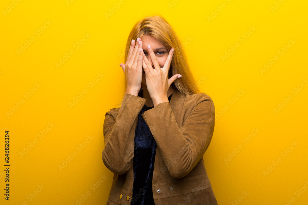 Blonde girl on vibrant yellow background covering eyes by hands and looking through the fingers Blonde girl on vibrant yellow background covering eyes by hands and looking through the fingers