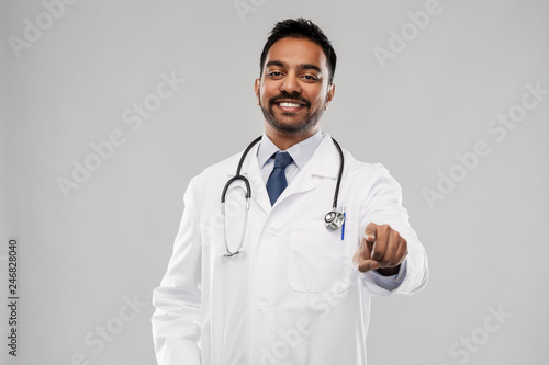medicine, profession and healthcare concept - smiling indian male doctor in white coat with stethoscope pointing finger to you over grey background
