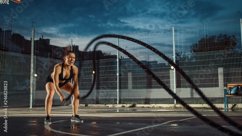Beautiful Energetic Fitness Girl Doing Exercises with Battle Ropes. She is Doing a Workout in a Fenced Outdoor Basketball Court and Shouting. Evening After Rain in a Residential Neighborhood Area.