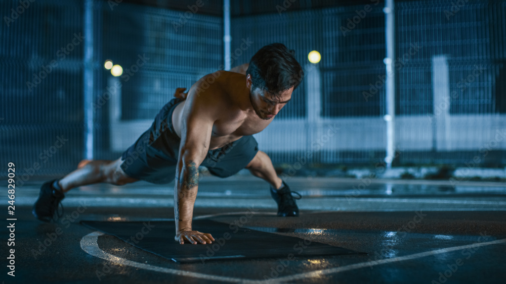 Strong Muscular Fit Shirtless Young Man is Doing One-Hand Push Up Exercises. He is Doing a Workout in a Fenced Outdoor Basketball Court. Night After Rain in a Residential Neighborhood Area. Strong Muscular Fit Shirtless Young Man is Doing One-Hand Push Up Exercises. He is Doing a Workout in a Fenced Outdoor Basketball Court. Night After Rain in a Residential Neighborhood Area.