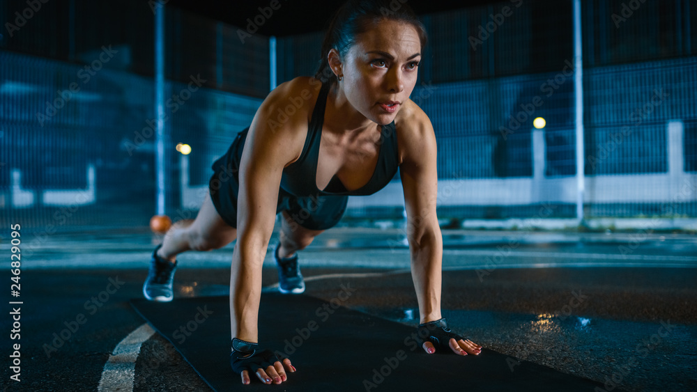 Fototapeta premium Beautiful Sporty Fitness Girl Doing Push Up Exercises. She is Doing a Workout in a Fenced Outdoor Basketball Court. Night Footage After Rain in a Residential Neighborhood Area.