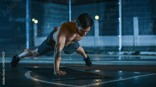 Strong Muscular Fit Shirtless Young Man is Doing One-Hand Push Up Exercises. He is Doing a Workout in a Fenced Outdoor Basketball Court. Night After Rain in a Residential Neighborhood Area.