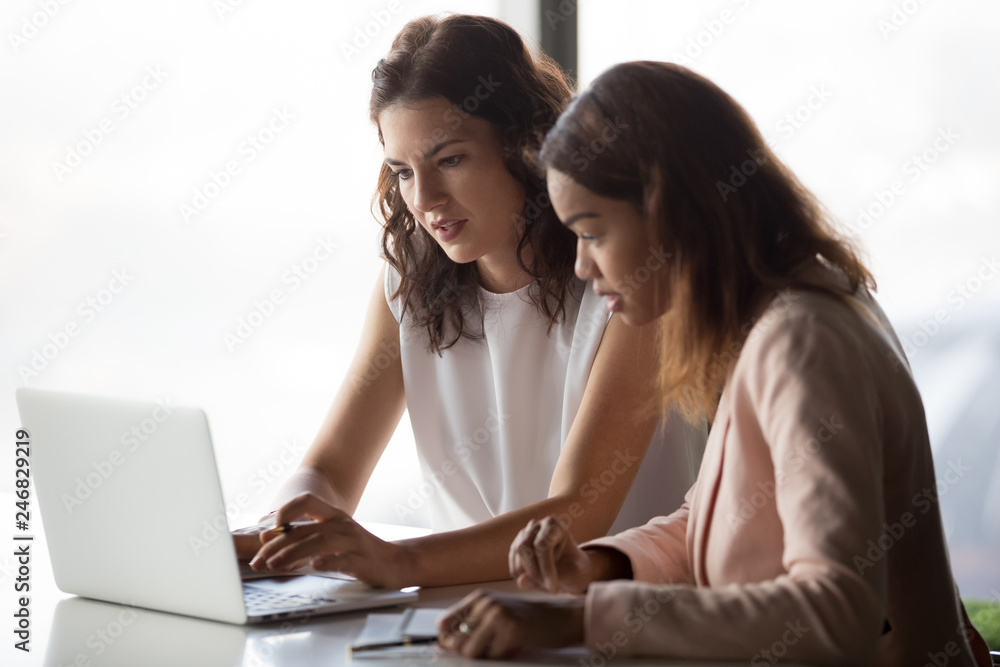 Serious diverse african and caucasian businesswomen workers talk discuss online project work together look at laptop in office, mentor teach intern, executive train employee using computer software Serious diverse african and caucasian businesswomen workers talk discuss online project work together look at laptop in office, mentor teach intern, executive train employee using computer software
