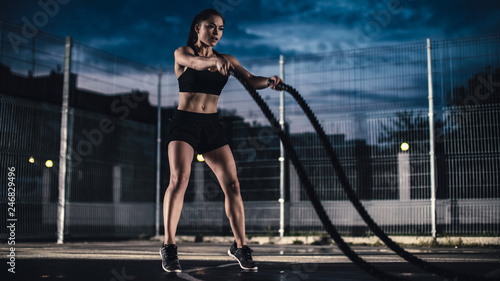 Beautiful Energetic Fitness Girl Doing Exercises with Battle Ropes. She is Doing a Workout in a Fenced Outdoor Basketball Court. Evening After Rain in a Residential Neighborhood Area.
