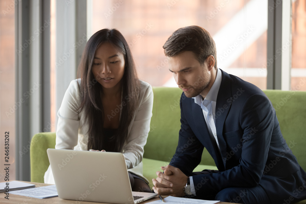 Serious focused asian businesswoman and caucasian businessman looking at laptop discussing online project results, making business offer to client or watching presentation using corporate software Serious focused asian businesswoman and caucasian businessman looking at laptop discussing online project results, making business offer to client or watching presentation using corporate software
