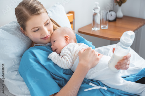 high angle view of smiling young mother holding baby bottle with milk and lying in bed with adorable baby