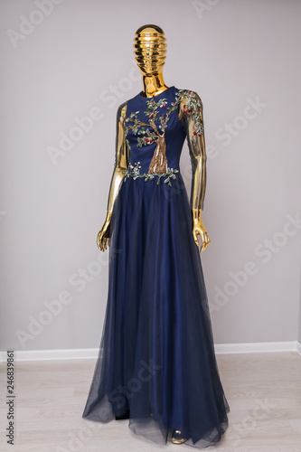 Long, blue designer, evening, women's dress handmade on gold, glossy mannequin. With white and gold ornament in the form of a cherry tree with fruits from beads. Оn gray background.
