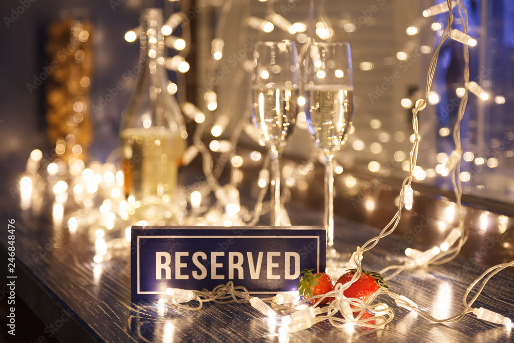 Closeup strawberry, two crystal glasses, bottle champagne sparkling wine, reserved nameplate on wooden table, window, garlands. Concept date evening in restaraunt, 8 march, Valentine's Day, christmas