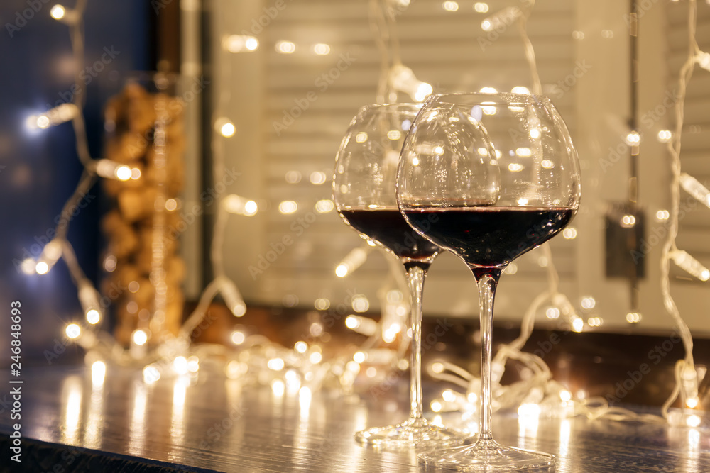 Closeup of two glasses of red wine in transparent crystal glasses on background of window, garlands. Concept of cozy evening in restaurant, date, christmas dinner, Valentine's Day Closeup of two glasses of red wine in transparent crystal glasses on background of window, garlands. Concept of cozy evening in restaurant, date, christmas dinner, Valentine's Day