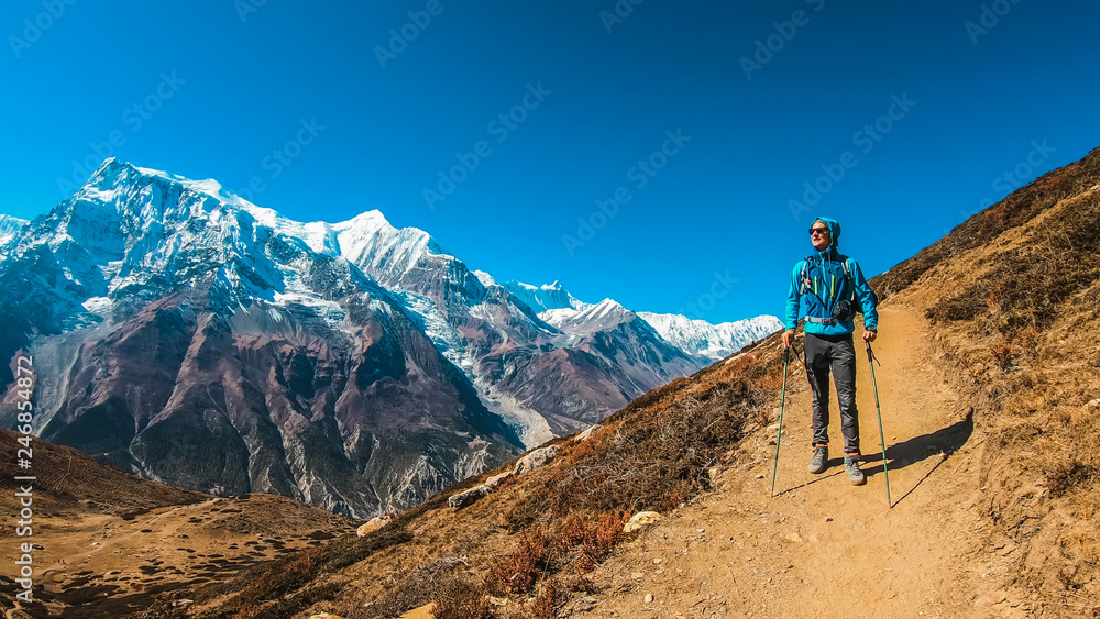 Trekking Young Man wears blue, the view on Annapurna Chain, Annapurna Circuit Trek, Nepal. Way to the Ice Lake. Man holds Nordic walking stick. Dry grass. Snowy mountains. Beautiful landscape