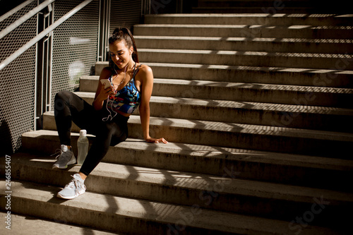 Young fit sporty woman resting and listen music on mobile phone after training outdoor on stairs in urban enviroment