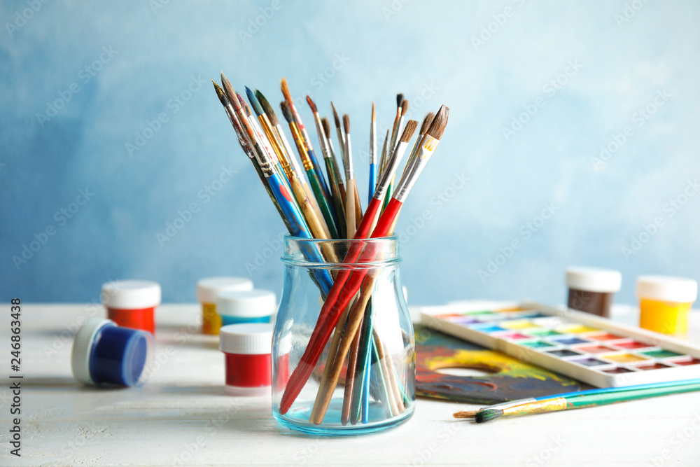 Fototapeta premium Glass jar with brushes and paints on table against color background