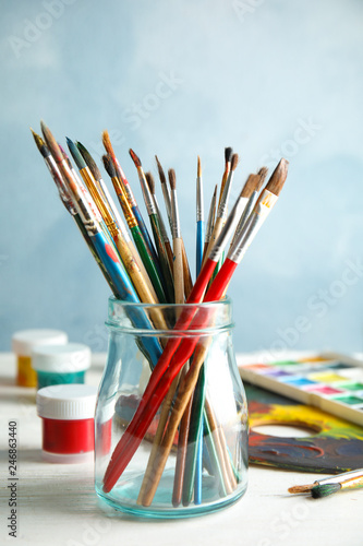 Glass jar with brushes and paints on table against color background