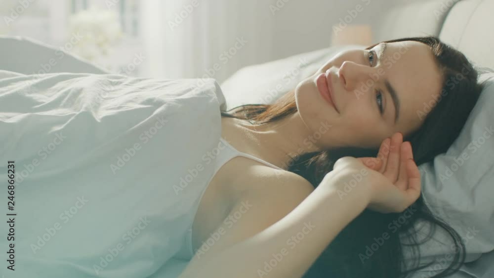 Beautiful Brunette Slowly Waking up in the Morning, Sits Up on the Bed, Smiles Charmingly. Sunrays Shine From a Window on a Happy Awaken Young Girl. Beautiful Brunette Slowly Waking up in the Morning, Sits Up on the Bed, Smiles Charmingly. Sunrays Shine From a Window on a Happy Awaken Young Girl.