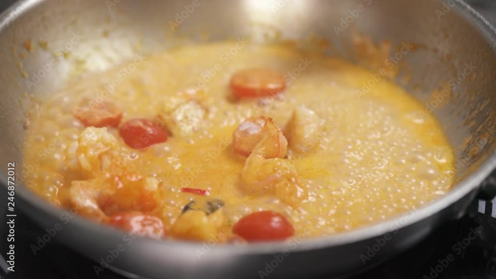 A top view of vegetables and shrimps sizzling in sauce on the frying pan and a hand adds some herbs, salt, mixes it with spoon and leaves it on the stove A top view of vegetables and shrimps sizzling in sauce on the frying pan and a hand adds some herbs, salt, mixes it with spoon and leaves it on the stove