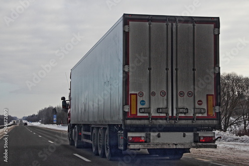 Logistics, international freight by road - dirty white european truck with semi trailer drive next to the two-lane asphalted highway road in the winter day, side rear view