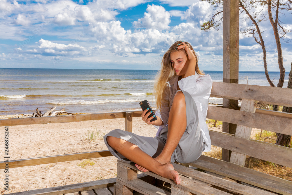Young girl with long fair hair sitting on a wooden pier on sea coast uses smartphone