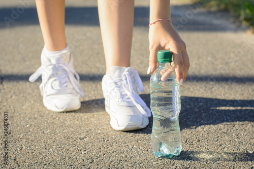 Young sportsmen girl in white sneakers stopped on the road to take a pause and drink some water out of plastic bottle during running in the morning in the countryside