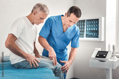 Canvas-taulu Dark-haired doctor wearing blue uniform examining knee