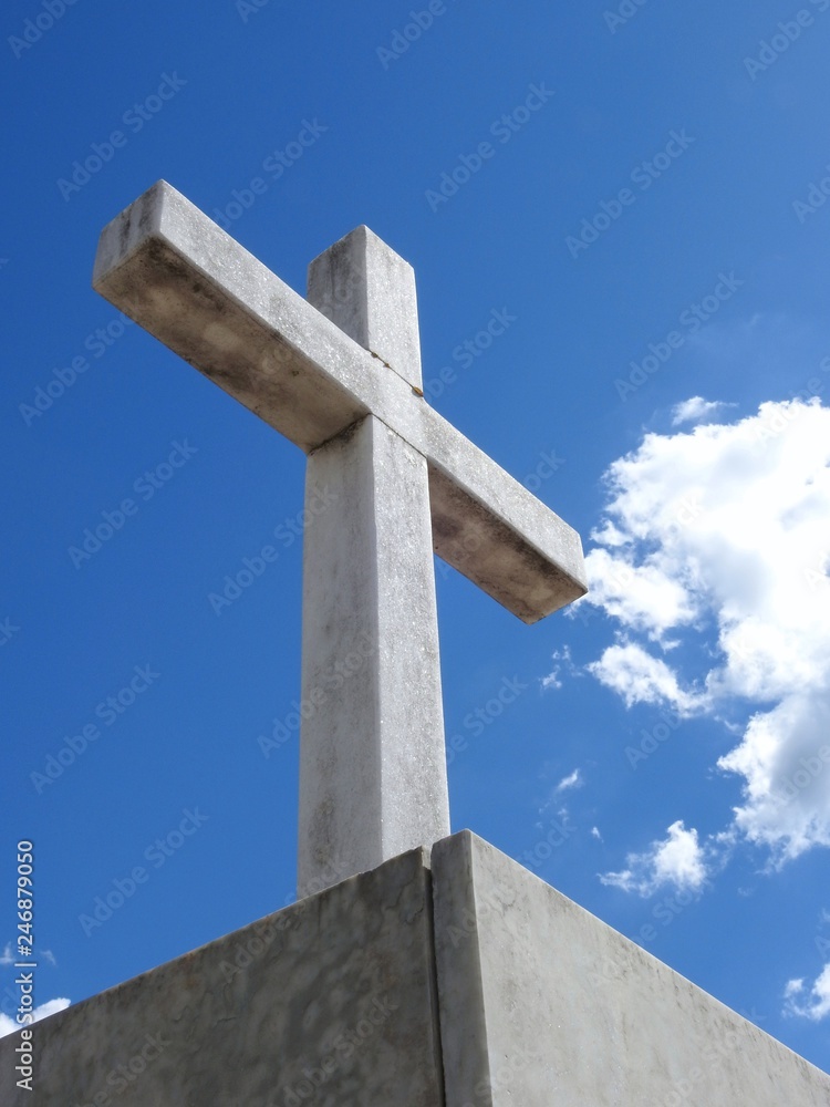 Scene in a graveyard: close-up of a religious stone cross illuminated by the sun. Blue sky with few clouds on a summer day. Bottom view.
