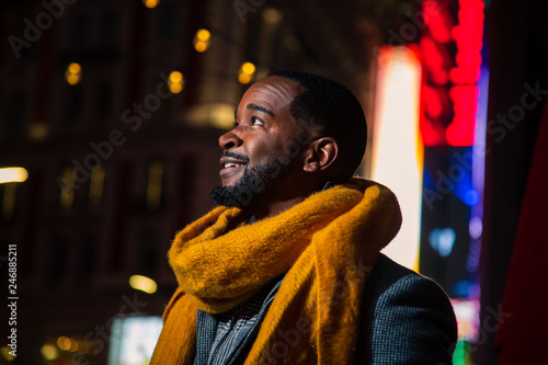 Black man in yellow scarf stands below a string of lights in an urban environment