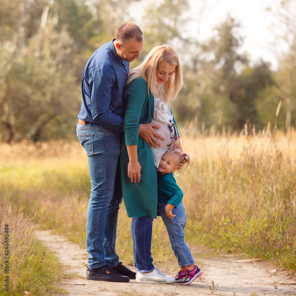 Fototapeta premium Portrait of pretty young pregnant woman with her young husband and cute little baby walking in park in sunny day. Family. Maternity. Happiness. Health.