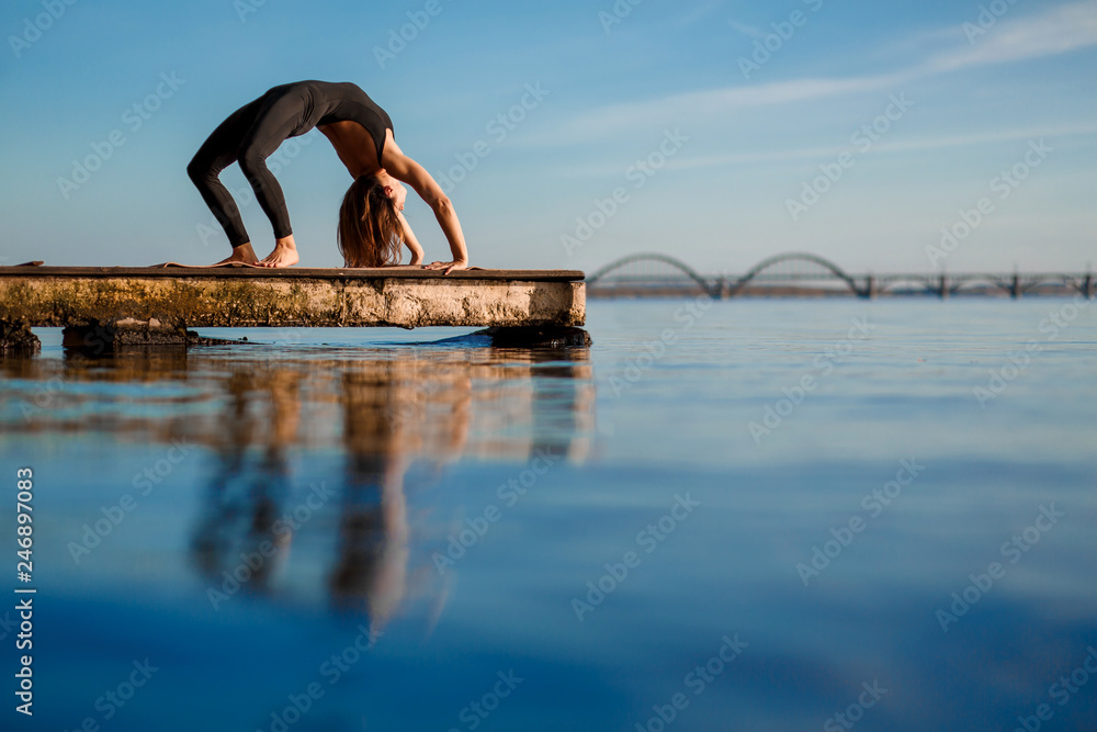 Fototapeta premium Young woman practicing yoga exercise at quiet wooden pier with city background. Sport and recreation in city rush
