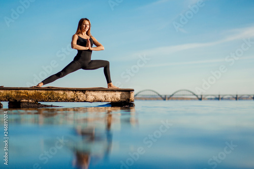 Young woman practicing yoga exercise at quiet wooden pier with city background. Sport and recreation in city rush