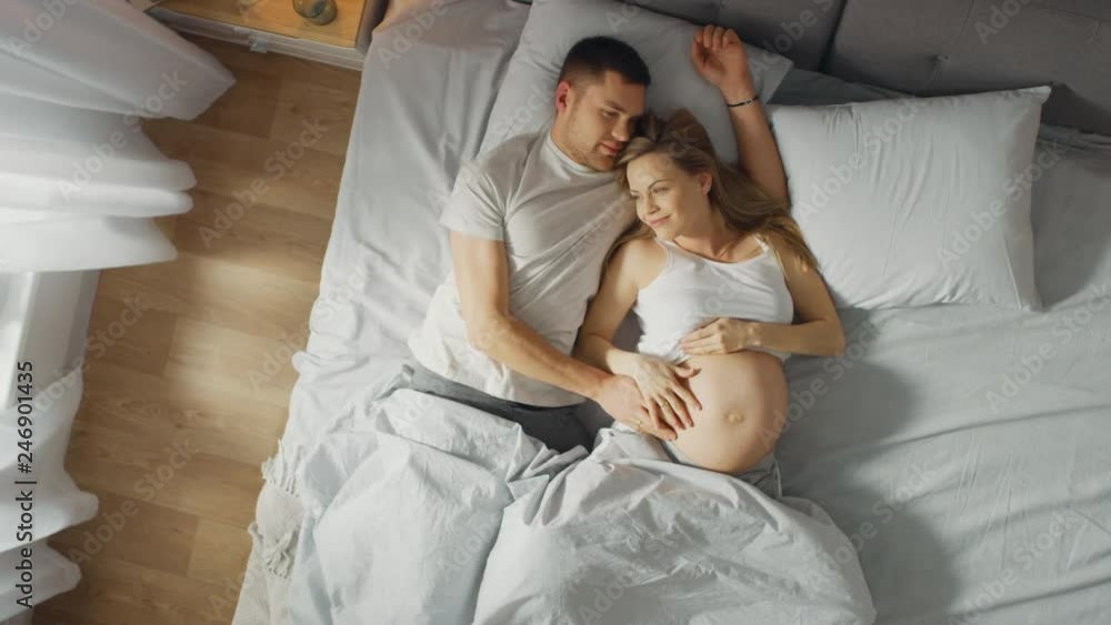 Happy Young Couple Cuddling Together in the Bed, Young Woman is Pregnant and Loving Partner Touches and Caresses Her Belly Tenderly. Top Down Camera. Happy Young Couple Cuddling Together in the Bed, Young Woman is Pregnant and Loving Partner Touches and Caresses Her Belly Tenderly. Top Down Camera.