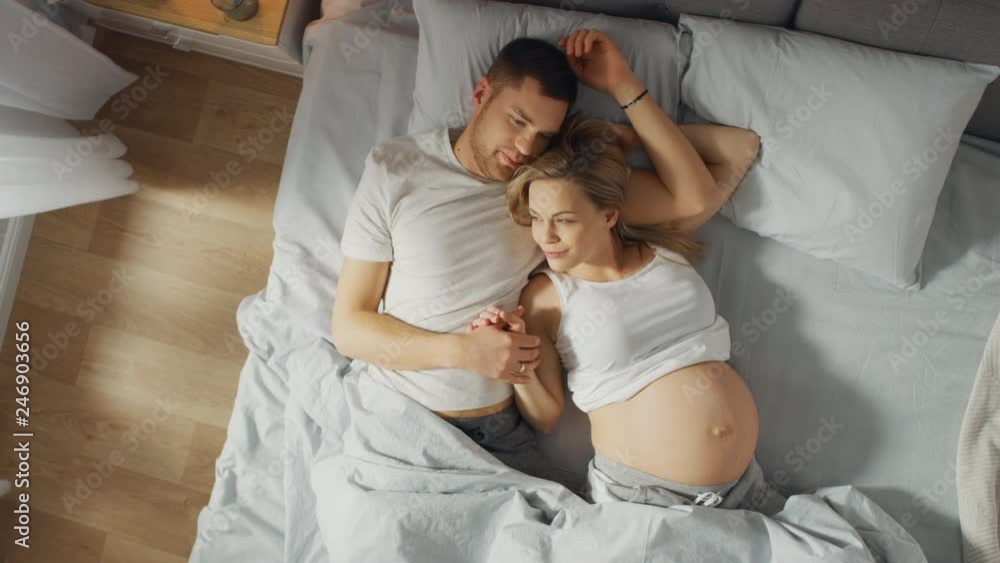 Happy Young Couple Cuddling Together in the Bed, Young Woman is Pregnant and Loving Partner Touches and Caresses Her Belly Tenderly. Sun Shines. Top Down Zoom out Camera Shot . Happy Young Couple Cuddling Together in the Bed, Young Woman is Pregnant and Loving Partner Touches and Caresses Her Belly Tenderly. Sun Shines. Top Down Zoom out Camera Shot .