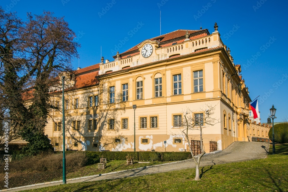 Prague, Czech Republic / Europe - January 31 2019: Former castle in Liben, Prague, now a seat of municipal authority, wall clock, yellow facade, red roof, Czech flag, paving, green grass, sunny day