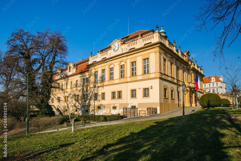 Obraz premium Prague, Czech Republic / Europe - January 31 2019: Former castle in Liben, Prague, now a seat of municipal authority, wall clock, yellow facade, red roof, Czech flag, paving, green grass, sunny day