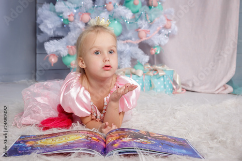 Portrait of cute european little blonde princess girl with crown in beautiful dress lying on the floor and reading book in decorated studio in christmas location with toys and christmas tree