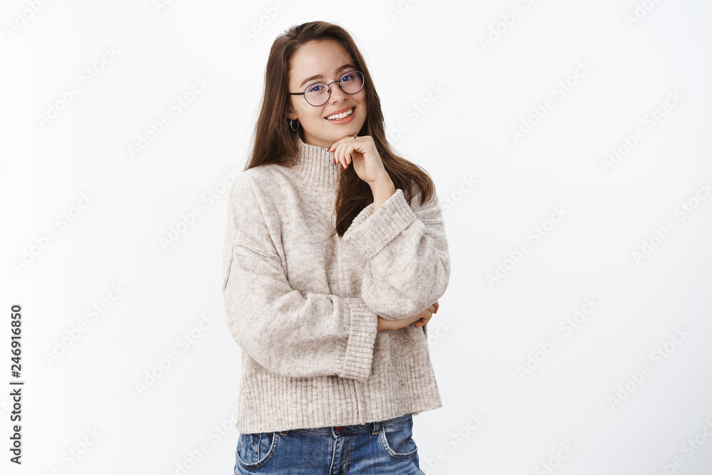 Waist-up shot of happy charismatic young brunette in glasses and warm sweater tilting head and smiling holding hand on chin, crossing arm against chest having conversation over gray background Waist-up shot of happy charismatic young brunette in glasses and warm sweater tilting head and smiling holding hand on chin, crossing arm against chest having conversation over gray background