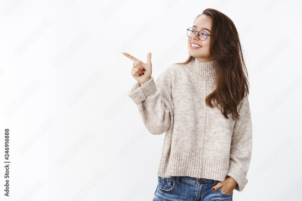 Curious and delighted cute girlfriend in sweater and glasses looking and pointing left, smiling broadly satisfied with interesting and funny exhibition, being intrigued by product showing in to friend