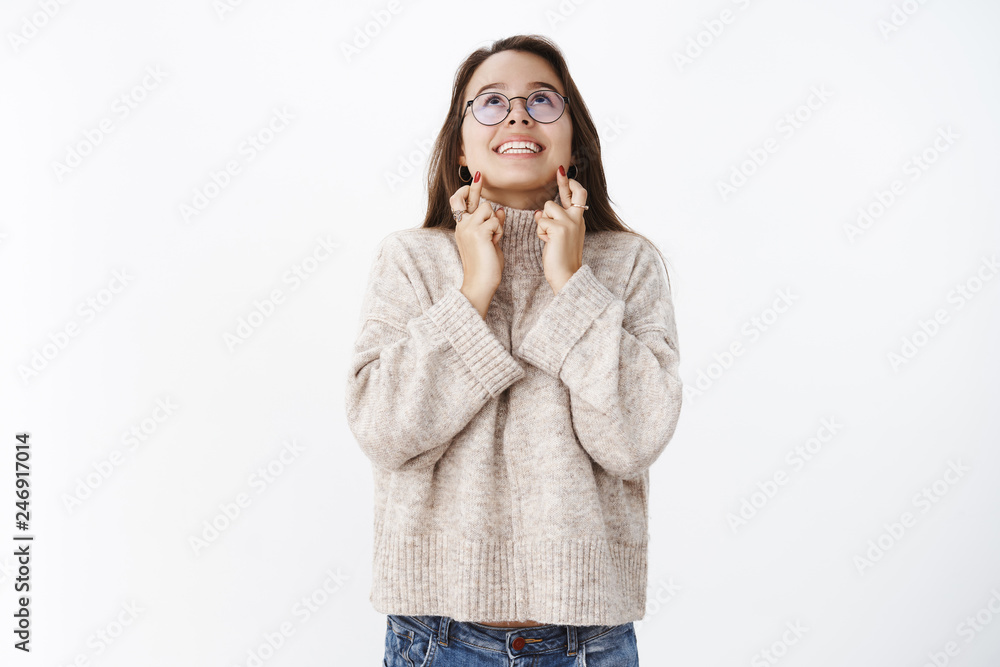Studio shot of thrilled and happy excited attractive young woman in glasses and sweater crossing fingers for good luck looking up with broad smile, praying, making wish asking god for dream come true Studio shot of thrilled and happy excited attractive young woman in glasses and sweater crossing fingers for good luck looking up with broad smile, praying, making wish asking god for dream come true