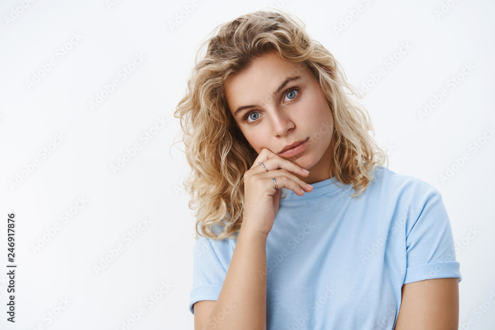Close-up shot of attractive feminine tanned young blond girl with blue eyes tilting head holding fingers near lip looking passionate and sincere at camera as listening over white background Close-up shot of attractive feminine tanned young blond girl with blue eyes tilting head holding fingers near lip looking passionate and sincere at camera as listening over white background