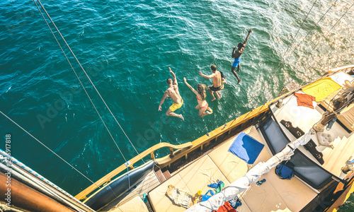 Aerial view of happy millenial friends jumping from sailboat on sea ocean trip - Rich guys and girls having fun together in exclusive boat party day - Luxury vacation concept on contrast bright filter