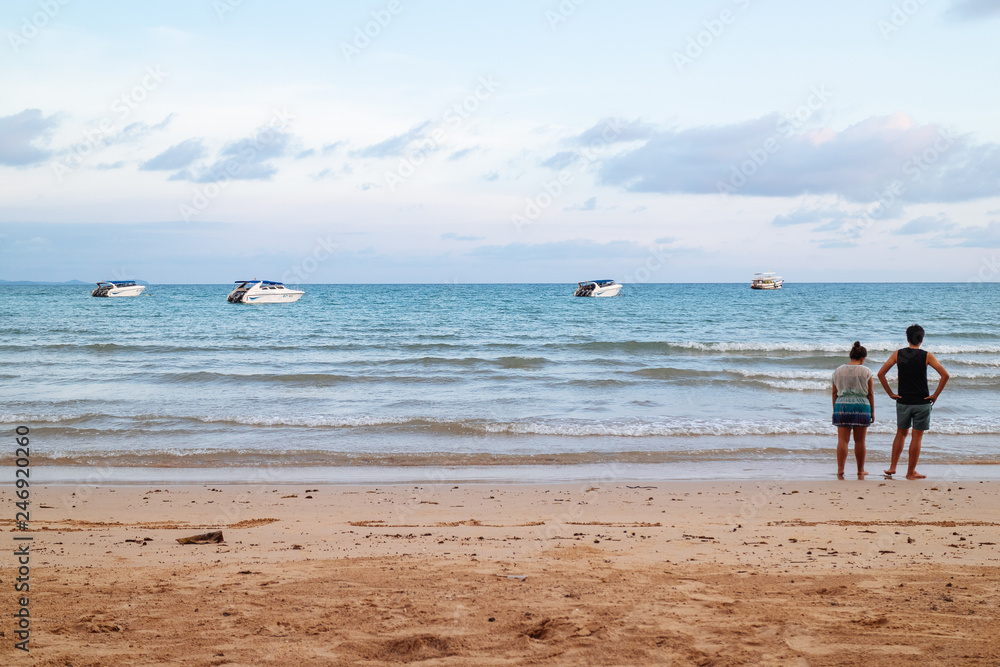 Back of a couple standing on the beach and looking to the sea with speed boats and bright sky in background at Koh Mak Island in Trat, Thailand.