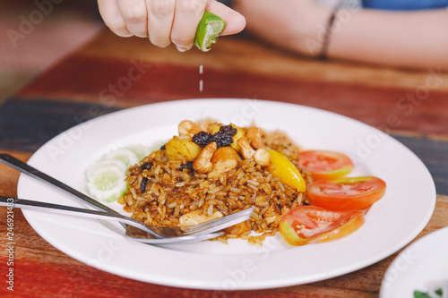 Close up Thai Pineapple Fried Rice topping with raisin, cashews and dried shredded pork. Served with cucumber and tomato and hand squeezing lime.