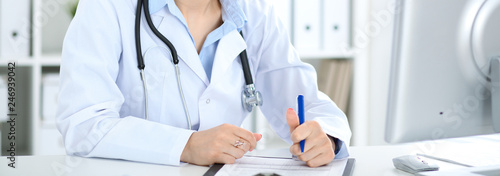 Unknown female doctor working at the desk with clipboard in hospital and filling up medical history form, close-up view. Medicine concept