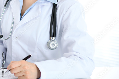 Unknown female doctor working at the desk with clipboard in hospital and filling up medical history form, close-up view. Medicine concept