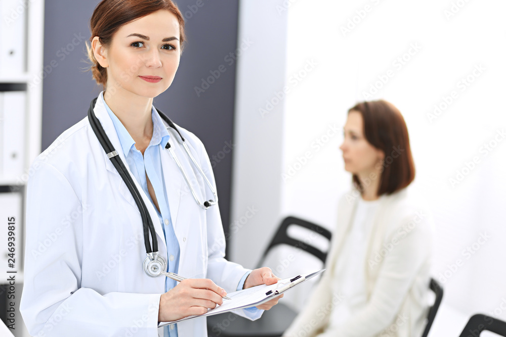 Doctor woman at work. Portrait of female physician filling up medical form while standing near reception desk at clinic or emergency hospital. Patient woman sitting at the background. Medicine concept