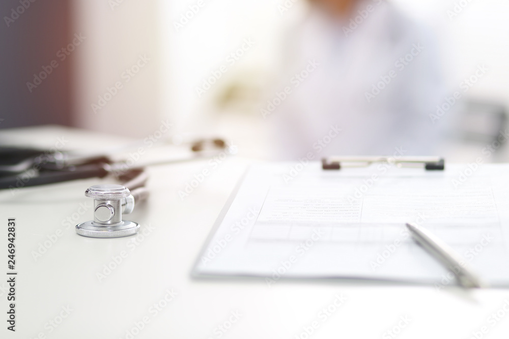 Stethoscope, prescription medical form lying on hospital reception desk with laptop computer and busy doctor at the background. Medical tools at doctor working table.Medicine concept Stethoscope, prescription medical form lying on hospital reception desk with laptop computer and busy doctor at the background. Medical tools at doctor working table.Medicine concept