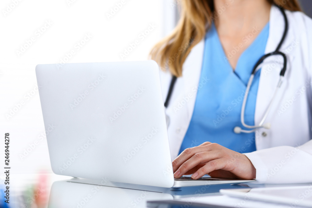 Doctor typing on laptop computer while sitting at the glass desk in hospital office. Physician at work. Medicine and healthcare concept Doctor typing on laptop computer while sitting at the glass desk in hospital office. Physician at work. Medicine and healthcare concept